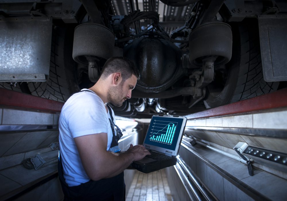 Vehicle mechanic with diagnostic tool laptop working under the truck in workshop.